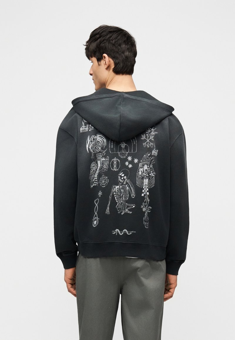 Νέος άνδρας με μαύρο φούτερ με κουκούλα, στο πίσω μέρος του οποίου υπάρχουν λευκά αφηρημένα σχέδια και σχέδια σκελετού, συνδυασμένο με γκρι παντελόνι, στέκεται μπροστά σε λευκό φόντο.