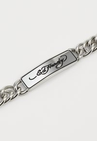 Bracelet en chaîne argentée avec une étiquette rectangulaire plate et polie, sur laquelle "Ed Hardy" est gravé dans une police cursive.