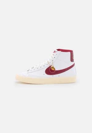 Biały sneaker za kostkę z czerwonym logo Nike, czerwonym kołnierzem wokół kostki, kremową podeszwą, białymi sznurowadłami i małą złotą zawieszką przymocowaną do sznurowadeł.