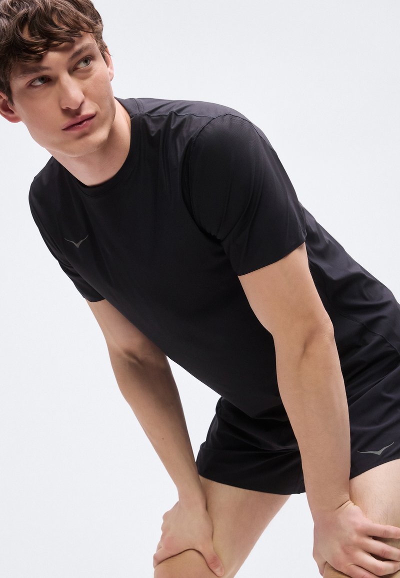 Giovane uomo in abbigliamento sportivo nero piegato in avanti con le mani sulle ginocchia, che guarda di lato su uno sfondo chiaro e uniforme.