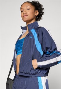 Chaqueta de deporte azul marino con detalles en azul claro y blanco, bolsillos con cremallera y puños elásticos, llevada sobre un sujetador deportivo de rayas azules.