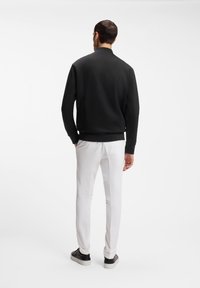 Άνδρας που φοράει μαύρο μακρυμάνικο πάνω μέρος, λευκό παντελόνι και μαύρα αθλητικά παπούτσια, στέκεται με τα χέρια στις τσέπες, γυρισμένος προς τα πίσω σε λευκό φόντο.