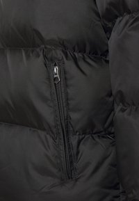 Schwarze Pufferjacke mit einer glatten, strukturierten Oberfläche. Verfügt über eine seitliche Tasche mit Reißverschluss und horizontale Steppnähte zur Isolation.