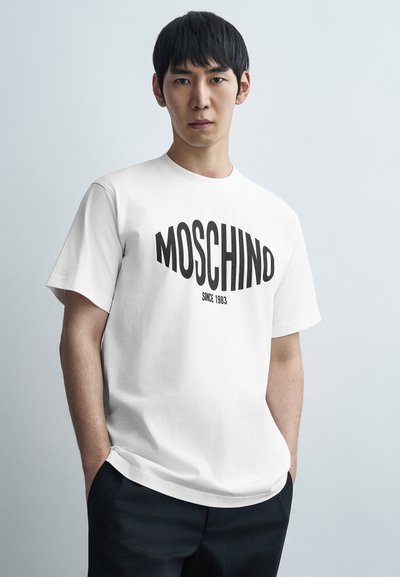 Λευκό βαμβακερό T-shirt με εντυπωσιακή μαύρη επιγραφή "MOSCHINO" και λεπτομέρεια "SINCE 1983". Στρογγυλή λαιμόκοψη και κοντά μανίκια. Ομαλή υφή.
