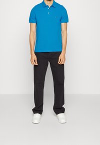 Muž v jasně modrém polokošili, černých rovně střižených džínách a bílých teniskách stojí proti jednoduchému pozadí.