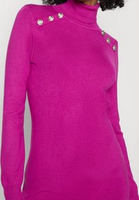 Pull col roulé ajusté en rose vif, texture côtelée, avec quatre boutons brillants sur l'épaule gauche. Matière douce et extensible.
