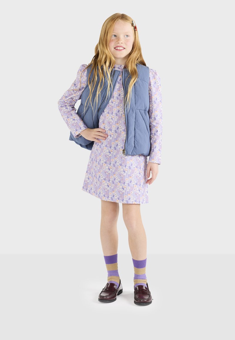 Blå quiltet vest over en pastel lilla blomsterkjole, parret med stribede sokker i lilla og gul, samt burgunder loafers.