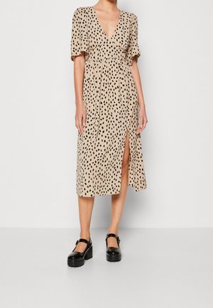 Beiges Midikleid mit schwarzem Punktmuster, V-Ausschnitt, kurzen Ärmeln und seitlichem Schlitz. Getragen mit schwarzen Plateauschuhen im Mary-Jane-Stil.