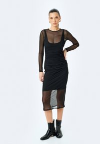 Czarna sukienka z połączeniem przezroczystej i nieprzezroczystej tkaniny, długimi rękawami oraz dopasowanym krojem. Marszczenia wzdłuż torsu, warstwowy dół.