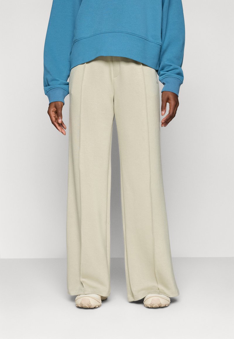 Pantaloni felpati beige a gamba larga realizzati in un tessuto morbido, con un discreto dettaglio di cucitura verticale. Abbinati a una felpa blu e calzature beige.