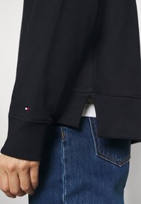 Felpa nera con polsini a coste, fessure laterali all'orlo e un piccolo dettaglio del logo rosso, bianco e blu sulla manica. Indossata con jeans blu.