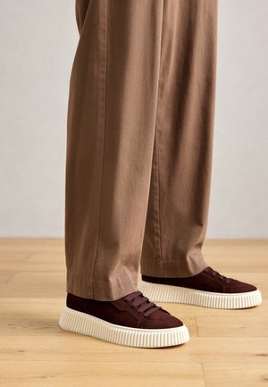 Person wearing brown wide-leg pants and dark brown sneakers with thick white ridged soles standing on wooden floor.