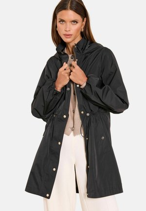 Woman wearing black long raincoat over beige cardigan and white pants, fastening coat zipper with both hands.