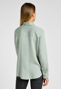 Chemisier vert clair à manches longues, avec col et poignets à bouton, fabriqué en tissu lisse. Dispose d'un ourlet incurvé et d'un détail de empiècement au dos.