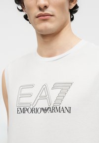 Νέος άνδρας που φοράει λευκή μπλούζα χωρίς μανίκια με μαύρο λογότυπο "EA7 Emporio Armani" τυπωμένο μπροστά, στραμμένος ελαφρώς προς τα αριστερά.