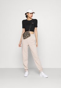 T-shirt nera corta con logo, pantaloni della tuta beige e sneakers bianche. Tiene una borsa a tracolla con stampa leopardata. Outfit casual con un'estetica sportiva.