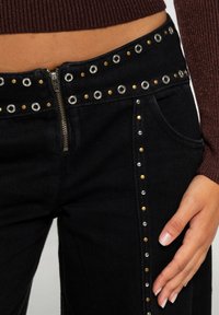 Pantalones vaqueros de mezclilla negra con cremallera frontal, decorados con ojales plateados y dorados a lo largo de la cintura y las costuras laterales.