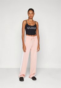 Melns crop tops ar "GUESS" logotipu, saskaņots ar gaismu rozā sporta biksēm ar baltām sānu panelēm. Modele valkā melnas slip-on kurpes.
