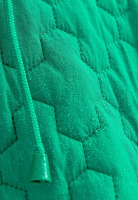 Tissu vert matelassé avec un motif hexagonal, présentant des gouttes d'eau à la surface et un cordon texturé avec une extrémité en plastique.