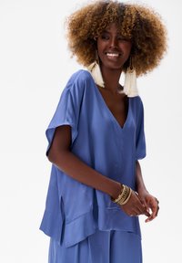 Luźna, krótka bluzka z miękkiego niebieskiego materiału, z dekoltem w kształcie litery V i bocznymi rozcięciami, z warstwowym dołem i gładką teksturą.