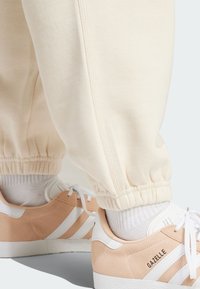 Krämfärgade joggers med elastiska manschetter, tillsammans med ljus persiko- och vita sneakers som har tre vita ränder och texturerade sulor.