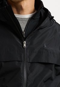 Musta kevyt takki, jossa on etuvetoketju, korkea kaulus ja pieni kirjailtu logo. Takissa on taskut sekä sileä, mattapintainen viimeistely.