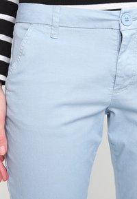 Ljusblå bomullsbyxor med platt midja, sidofickor och enstaka knappstängning. Slät textur med minimal detaljering.