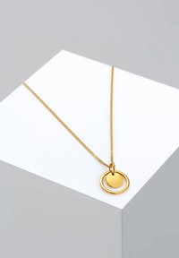 Collier doré comportant une chaîne fine et deux pendentifs circulaires entrelacés, l'un plat et l'autre en relief, sur une surface d'exposition blanche.