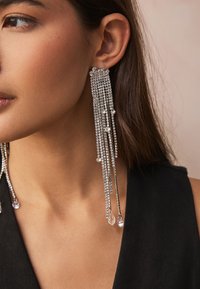 Long, silver earrings with cascading chains adorned with small clear gemstones, featuring a shiny and reflective surface.