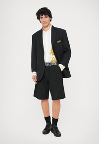 Μαύρο σακάκι πάνω από άσπρο πουκάμισο με χρυσό σχέδιο, σε συνδυασμό με μαύρες βερμούδες και μαύρες αθλητικές εσπαντρίγιες. Διαθέτει λογότυπο στην ζώνη και υφασμάτινες μανσέτες.