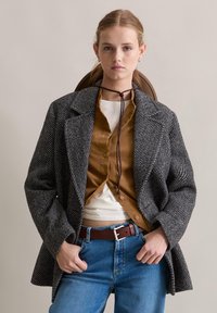Szary blazer w wzór jodełki z wełny z szerokimi klapami, noszony na brązowej koszuli z guzikiem i białej bluzce, w zestawie z niebieskimi jeansami z wysokim stanem.
