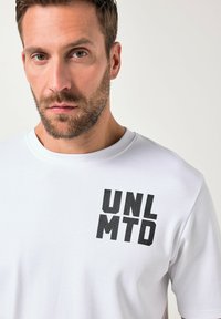Camiseta de algodón blanca con mangas cortas y cuello redondo, que presenta un llamativo texto en negro "UNL MTD" en la parte superior izquierda.