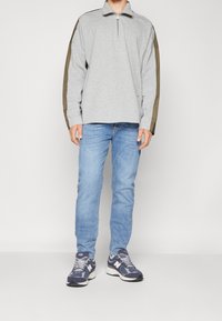 Sudadera gris de manga larga con cremallera y detalles en verde oliva, combinada con jeans azul claro y zapatillas navy con detalles en blanco.