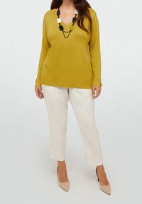 Donna che indossa un maglione a V giallo senape, pantaloni bianchi a vita alta, tacchi beige a punta e una collana geometrica in bianco e nero.
