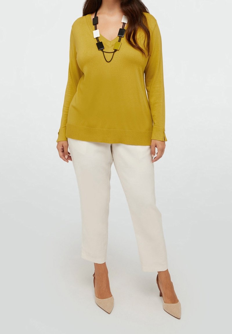 Donna che indossa un maglione a V giallo senape, pantaloni bianchi a vita alta, tacchi beige a punta e una collana geometrica in bianco e nero.