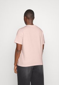 Tričko s krátkým rukávem, světle růžové barvy, vyrobené z bavlny, s kulatým výstřihem a volnějším střihem, v kombinaci s tmavě šedými kalhotami.