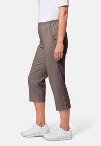 Leichte, braune cropped Hose mit elastischem Bund, Seitentaschen und drei dekorativen Knöpfen am Saum. Getragen mit weißen Sneakern.