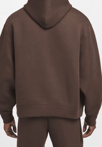 Haut à capuche marron en tissu doux et texturé. Dispose d'une grande capuche, d'épaules tombantes et de coutures simples. Pas de marquage ni d'accents visibles.