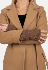 Abrigo de lana beige con solapas con muesca y bolsillos, superpuesto a mangas de punto marrones sin dedos; tejido texturizado, diseño cálido, corte casual.