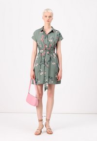 Abito camicia verde floreale con maniche corte, vita cintrata e un nodo laterale. Abbinato a una piccola borsa rosa e sandali beige con cinturini.