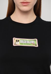Juodas marškinėlis su spalvingu grafiniu spaudiniu, primenančiu užkandžių pakuotę, su tekstu "1986 Moschino Jeans Suteikia Energijos" žaismingais šriftais.