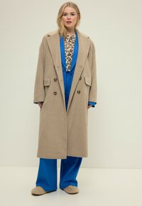 Abrigo de lana mezcla beige de doble botonadura con grandes botones, usado sobre un traje azul y una blusa estampada; persona de pie contra un fondo neutro.