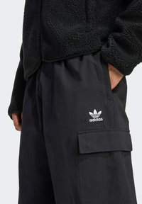 Chaqueta de forro polar negra con un acabado texturizado junto a pantalones negros con un bolsillo tipo cargador y el logo blanco de adidas. Diseño simple y funcional.