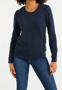 Marinblå långärmad tröja med texturerad tyg, rund halsringning och åtsittande mudd, tillsammans med blå jeans. Inga synliga mönster eller accentuerande detaljer.