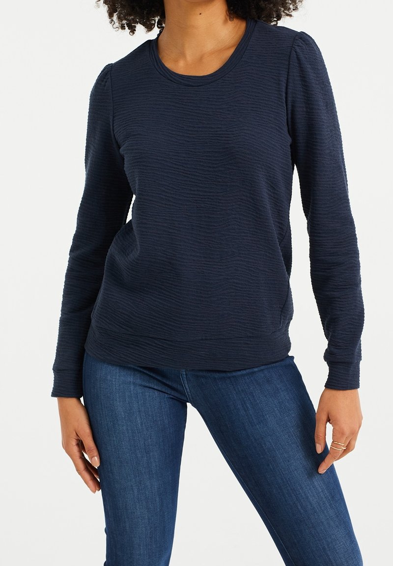 Marinblå långärmad tröja med texturerad tyg, rund halsringning och åtsittande mudd, tillsammans med blå jeans. Inga synliga mönster eller accentuerande detaljer.