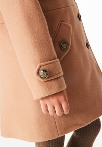 Abrigo de lana color beige claro con un acabado texturizado, que presenta grandes botones dorados y un detalle de puño entallado.