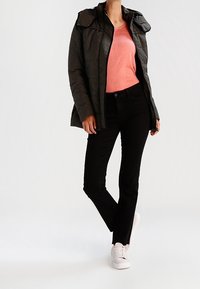 Dunkelgrüne gepolsterte Jacke mit Kapuze, kombiniert mit einem korallenfarbenen Oberteil, schwarzen Slim-Fit-Hosen und weißen Sneakern.