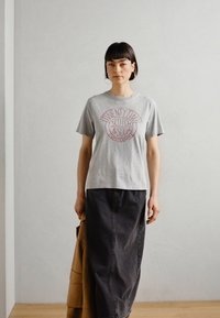 Grau meliertes T-Shirt mit Rundhalsausschnitt und einem kreisförmigen roten Grafikdruck mit der Aufschrift "TWENTY TWO SCOTCH & SODA", kombiniert mit einem langen dunklen Rock. Die Textur wirkt weich.