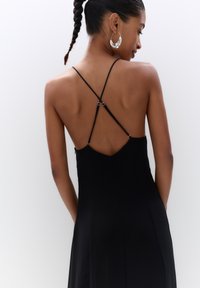 Schwarzes Kleid mit dünnen, verstellbaren Trägern und einem Kreuzdesign auf der Rückseite. Glatte Textur und eine taillierte Silhouette. Silberne Hardware-Akzente.