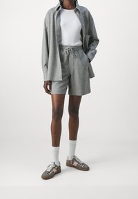 Camicia grigia a maniche lunghe con bottoni, abbinata a pantaloni corti grigi. T-shirt bianca sotto e sneakers grigio-marroni con accenti rosa.